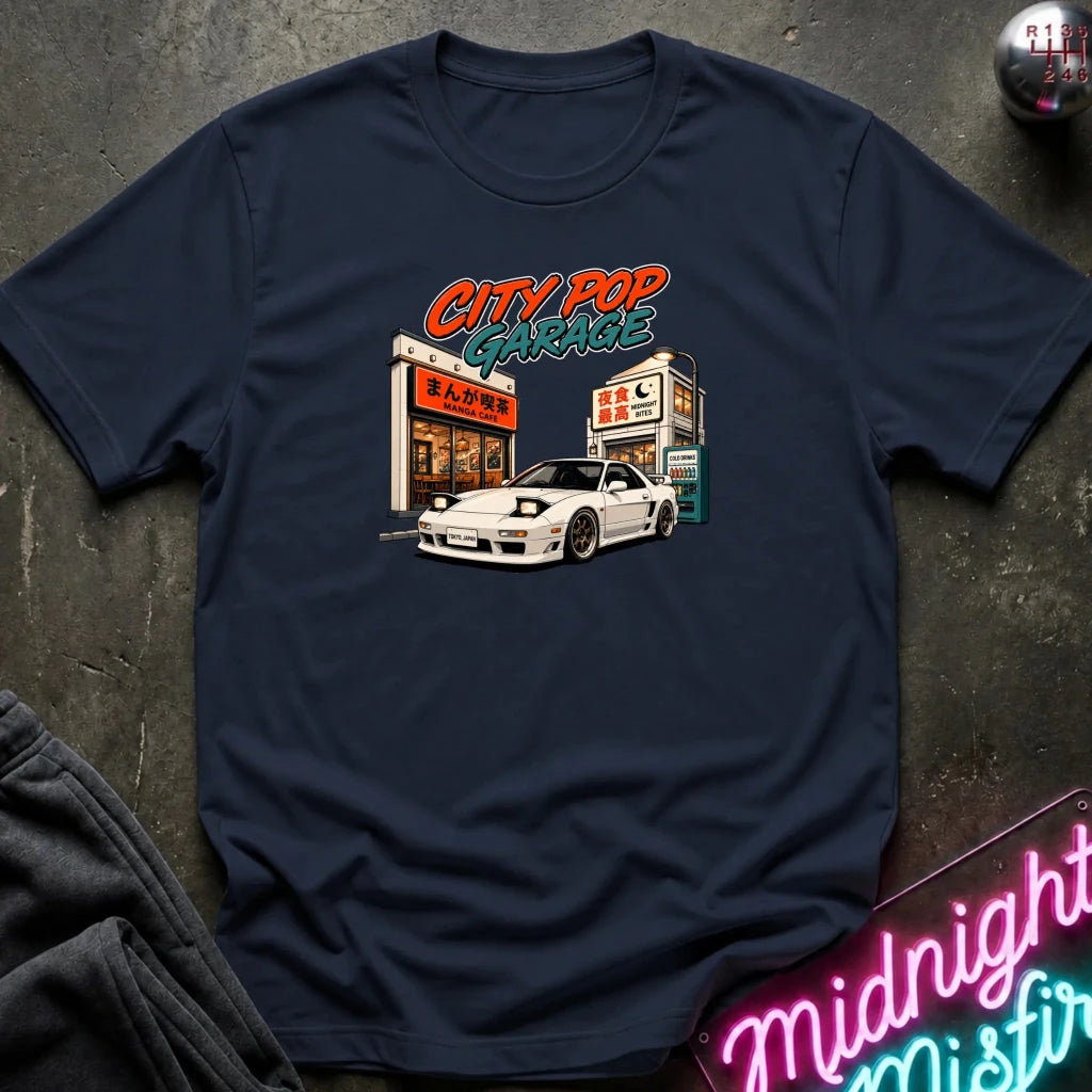 A navy blue graphic t-shirt laid flat on a dark textured surface. The shirt features a retro-style graphic of a white sports car parked in front of a Japanese-themed garage with the text 'CITY POP GARAGE' in orange and green.