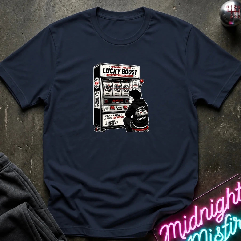 A navy blue short-sleeve t-shirt laid flat on a dark textured surface, featuring a graphic print of a person sitting in front of a slot machine with the text 'LUCKY BOOST'.