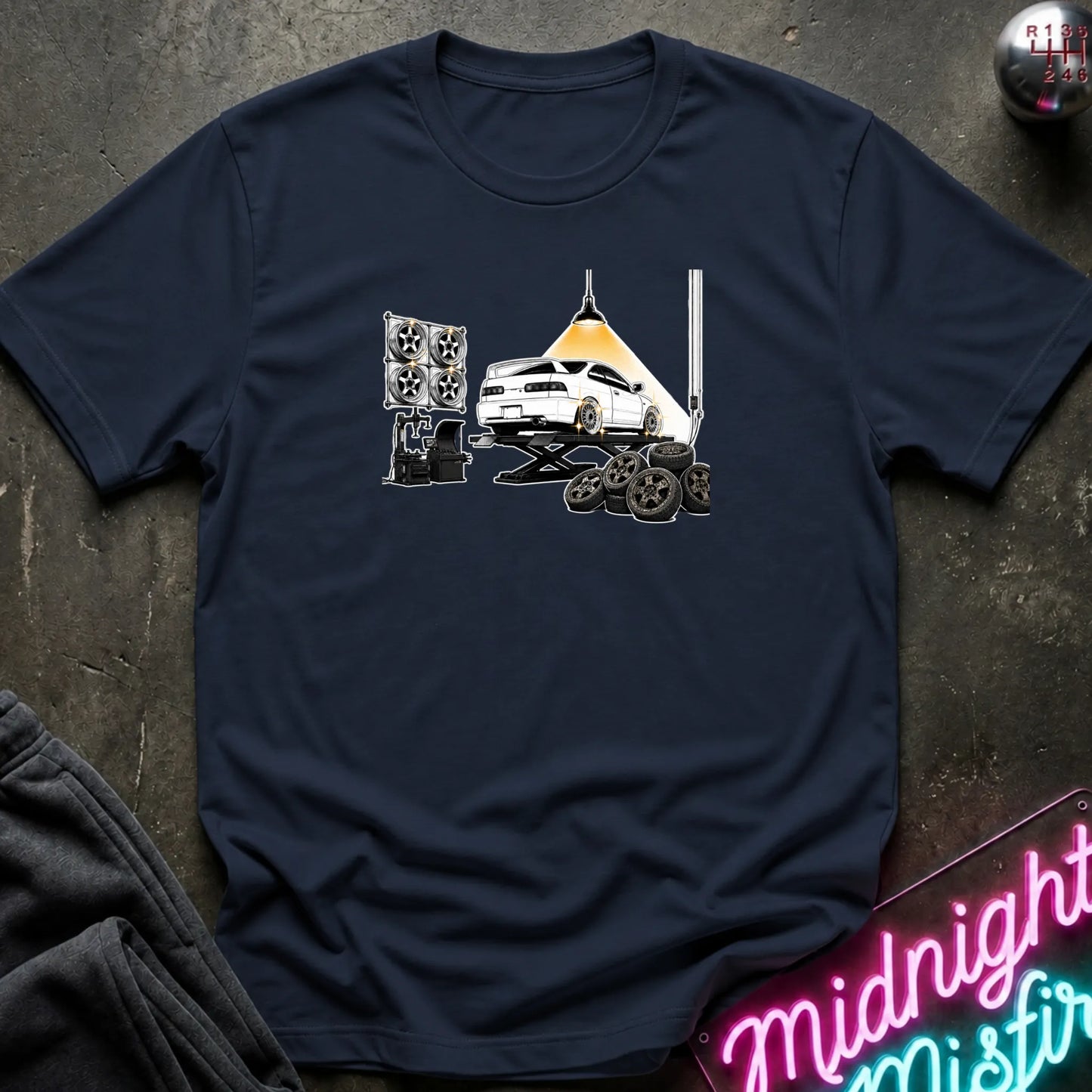 A navy blue short-sleeve crewneck t-shirt laid flat on a dark textured surface. The shirt features a graphic print of a white car on a hydraulic lift with new wheels.