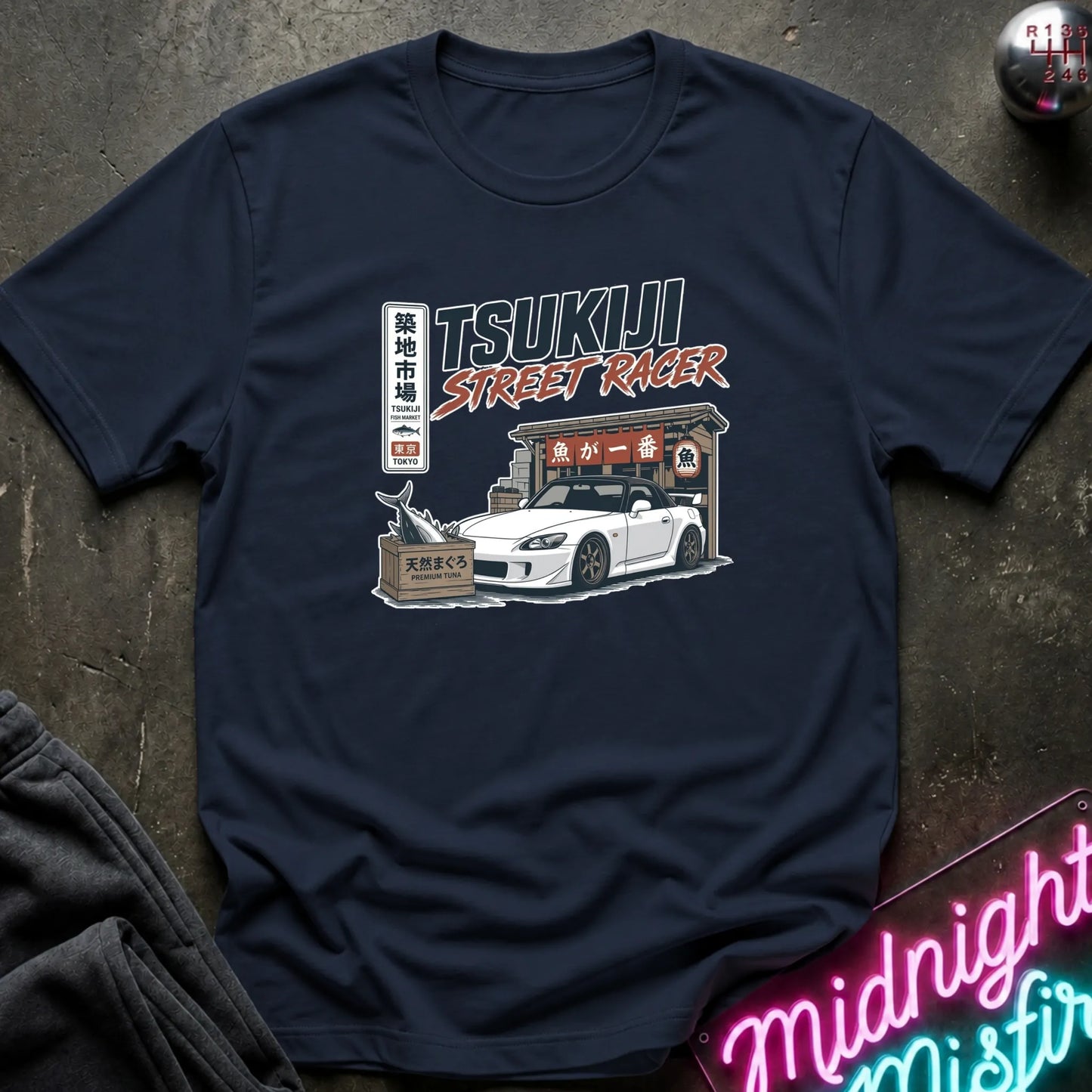 Navy short-sleeve graphic T-shirt laid flat on and surrounding an illustration of a white sports car parked outside a fish market.