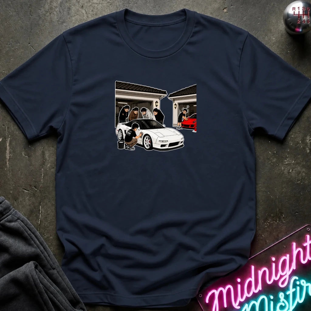 A navy blue short-sleeve t-shirt laid flat on a dark textured surface, featuring a central graphic print of a white sports car in front of a house with cartoon-style figures. 