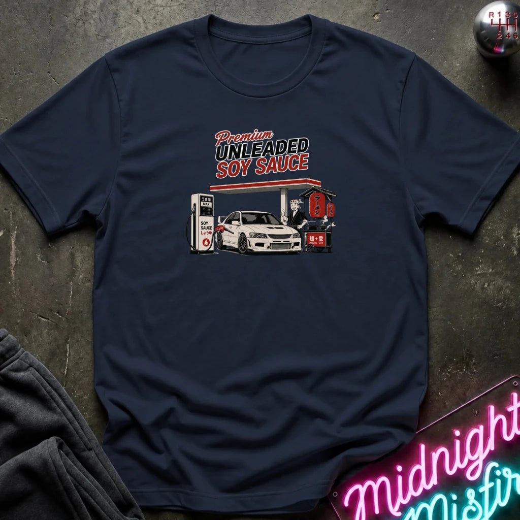A navy blue short-sleeve t-shirt with a retro-style graphic print reading 'Premium UNLEADED SOY SAUCE' above an illustration of a white sports car at a gas station, laid flat on a dark textured surface.
