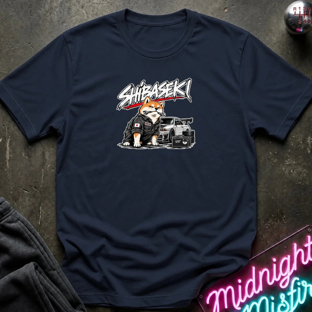 A navy blue crewneck t-shirt laid flat on a dark textured surface. The shirt features a graphic print of a Shiba Inu (shiba dog) wearing a black racing suit next to a black sports car, with the word 'SHIBASEKI' written above in a stylized white and red font.