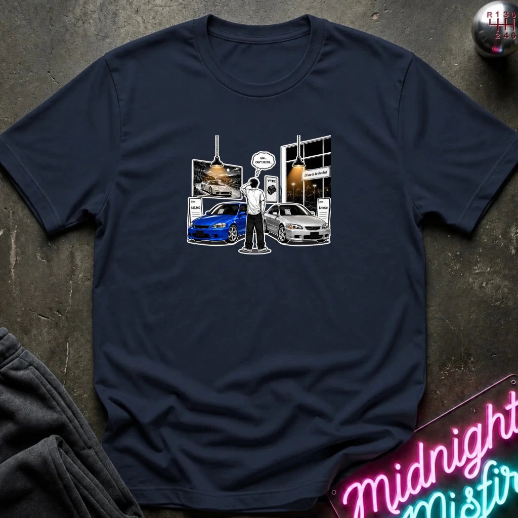 A navy blue short-sleeve t-shirt with a graphic print of a person holding a speedometer in front of a car dealership, featuring a blue sports car and a silver car. The shirt is laid flat on a dark textured surface with a neon sign and folded sweatpants nearby.