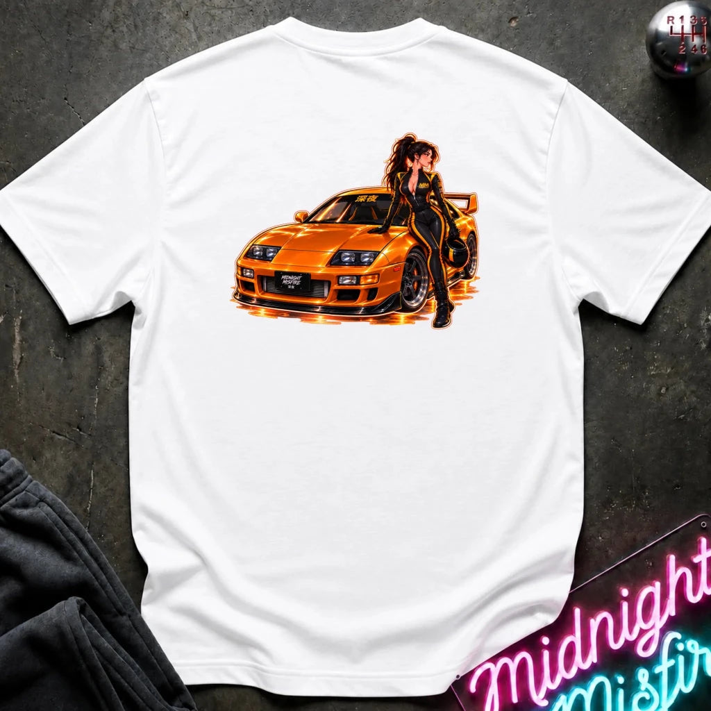 A white short-sleeve t-shirt laid flat on a dark textured surface, featuring a large graphic print of an orange sports car with a woman in a black outfit leaning on it. The shirt is styled with a dark sweatshirt in the corner and a neon sign in the background.
