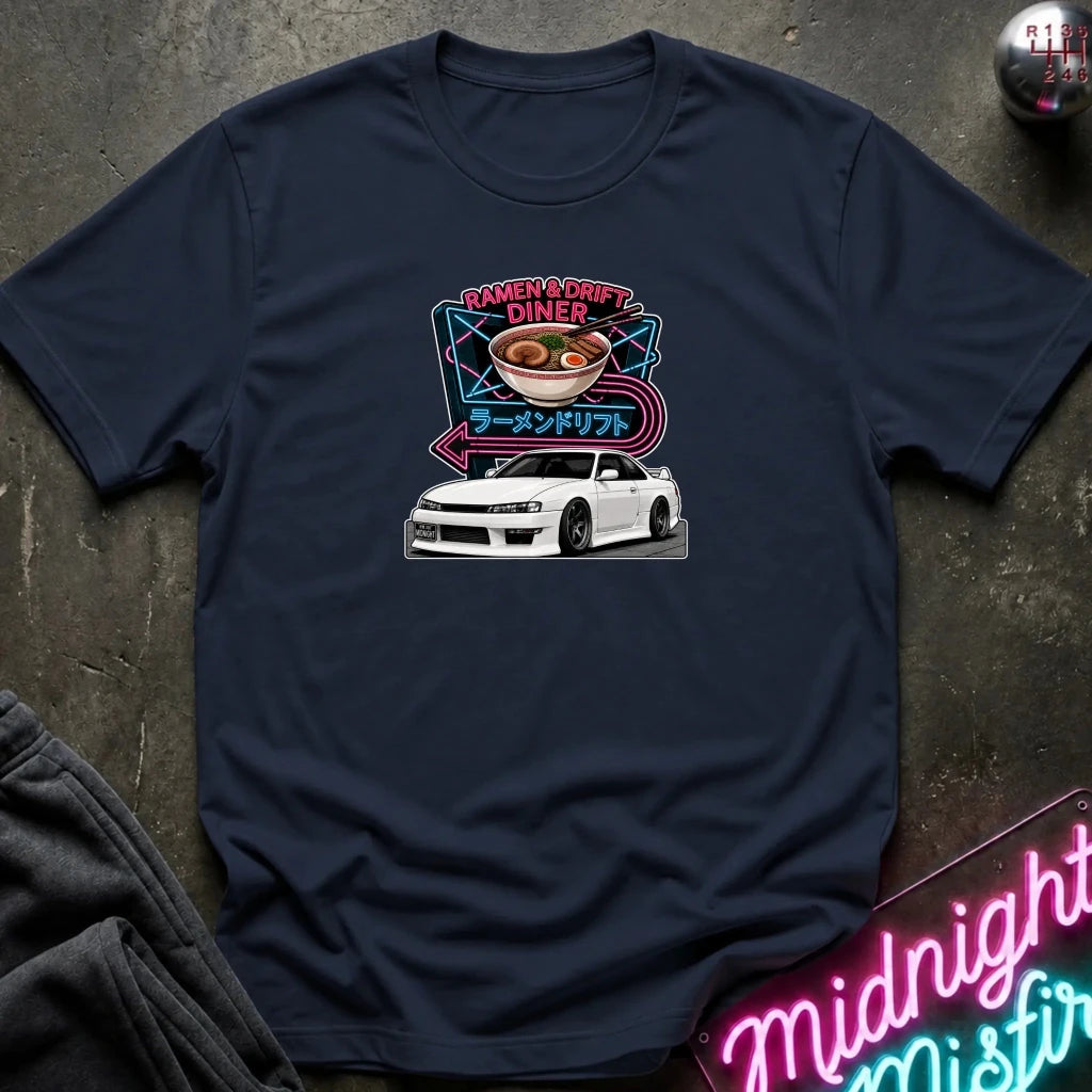 A navy blue crewneck t-shirt laid flat on a dark textured surface. The shirt features a graphic print of a white sports car under a neon sign that reads 'Ramen & Drift Diner' with Japanese text.