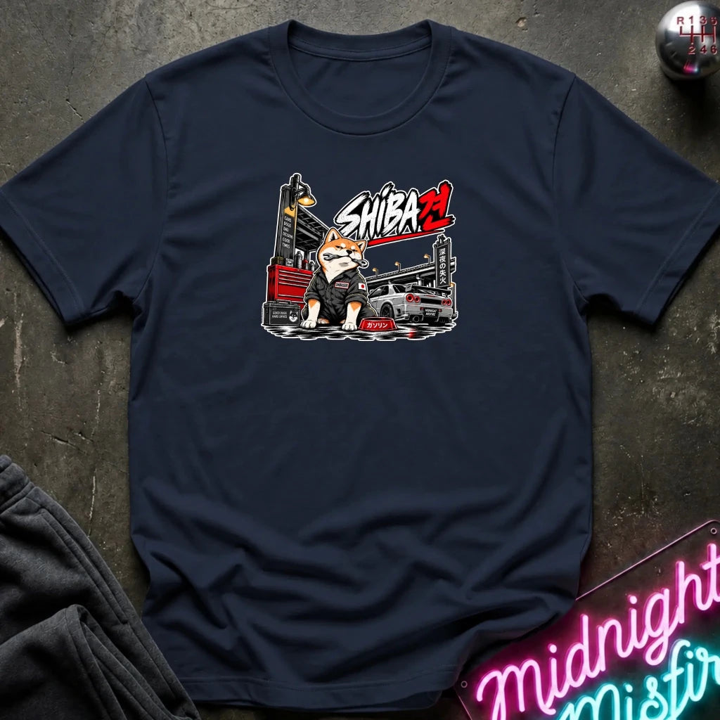 A navy blue graphic t-shirt laid flat on a dark textured surface. The graphic features a cartoon Shiba Inu character with a crowbar and a garage backdrop, with the text 'SHIBA' in red and white.