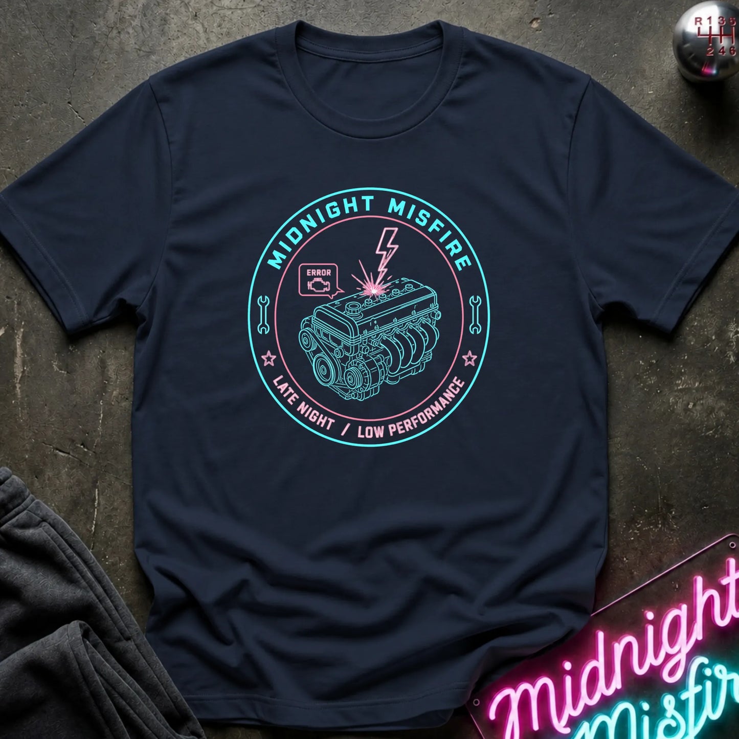 A navy blue short-sleeve t-shirt with a circular graphic on the chest depicting a car engine and the text 'MIDNIGHT MISFIRE' and 'LATE NIGHT / LOW PERFORMANCE' in teal and pink. The shirt is laid flat on a dark textured surface with a neon sign and dark fabric partially visible in the corners.