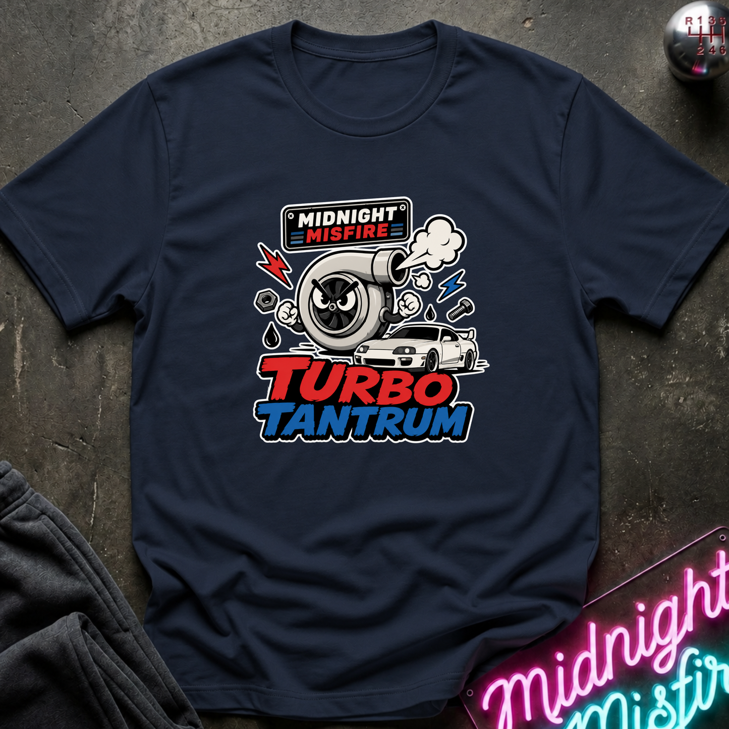 A navy blue short-sleeve t-shirt laid flat on a dark textured surface, featuring a colorful graphic print of a turbocharger, a white race car, and the text 'MIDNIGHT MISFIRE' and 'TURBO TANTRUM'.