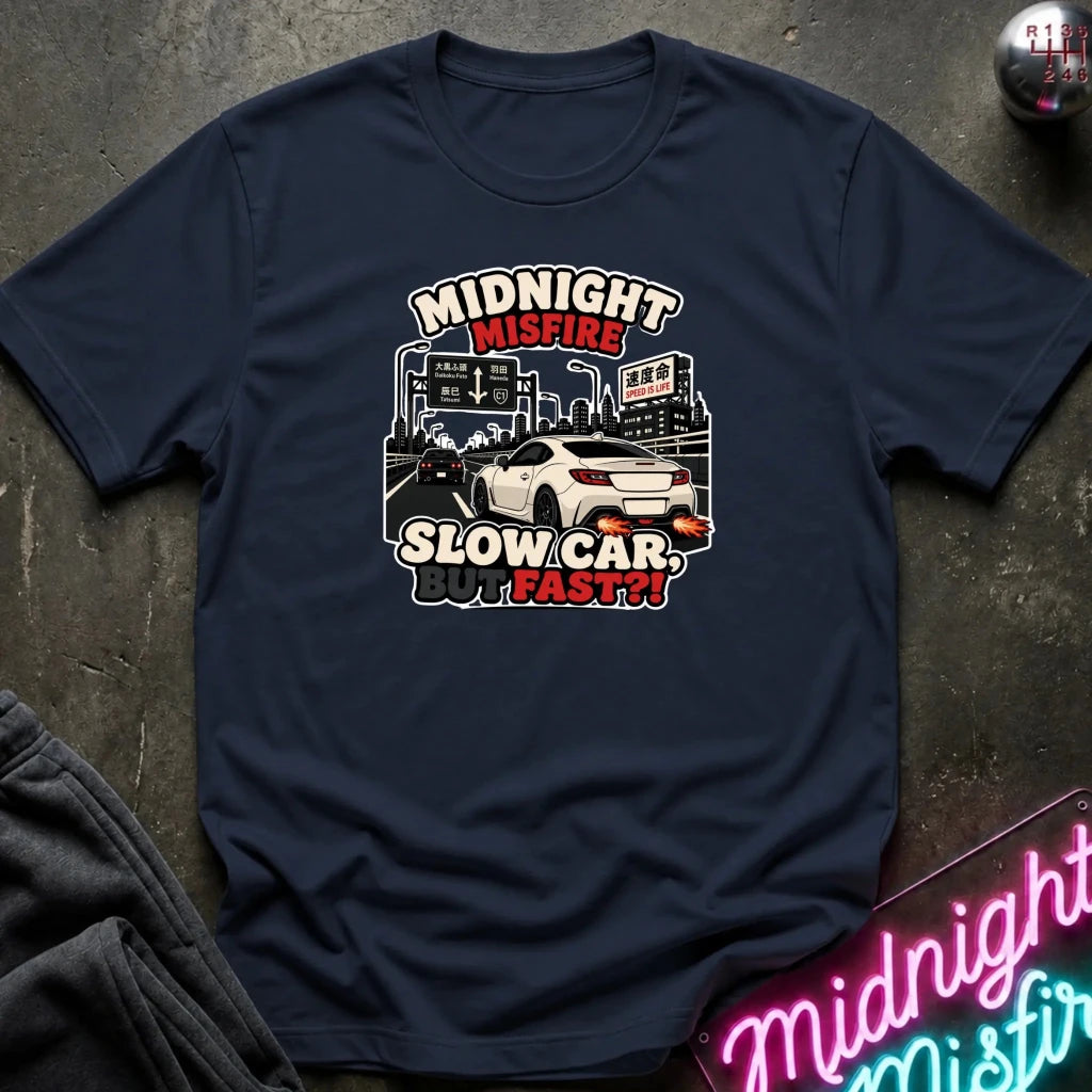 A navy blue graphic t-shirt laid flat on a dark textured surface. The graphic features a white sports car and the text 'MIDNIGHT MISFIRE SLOW CAR FAST!!' in stylized lettering.