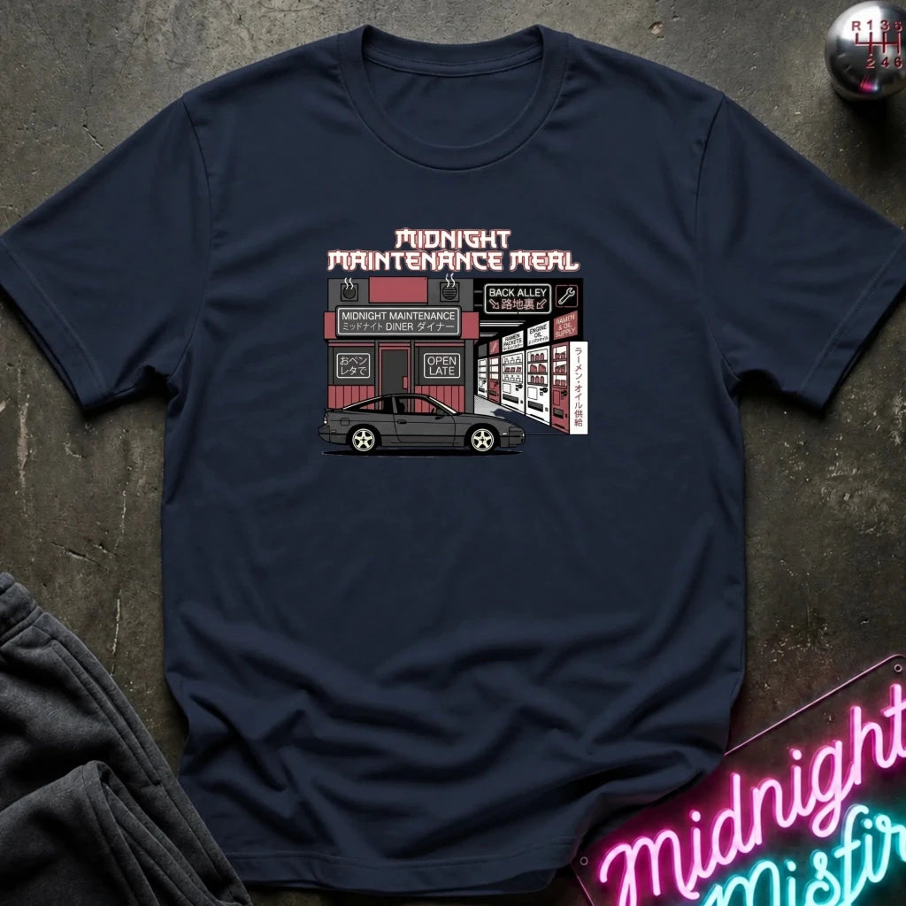 A navy blue crewneck t-shirt featuring a graphic print of a car repair shop scene with the text 'MIDNIGHT MAINTENANCE MEAL' in pink and white. The shirt is displayed flat on a dark textured surface with a neon sign and dark clothing partially visible in the corners.