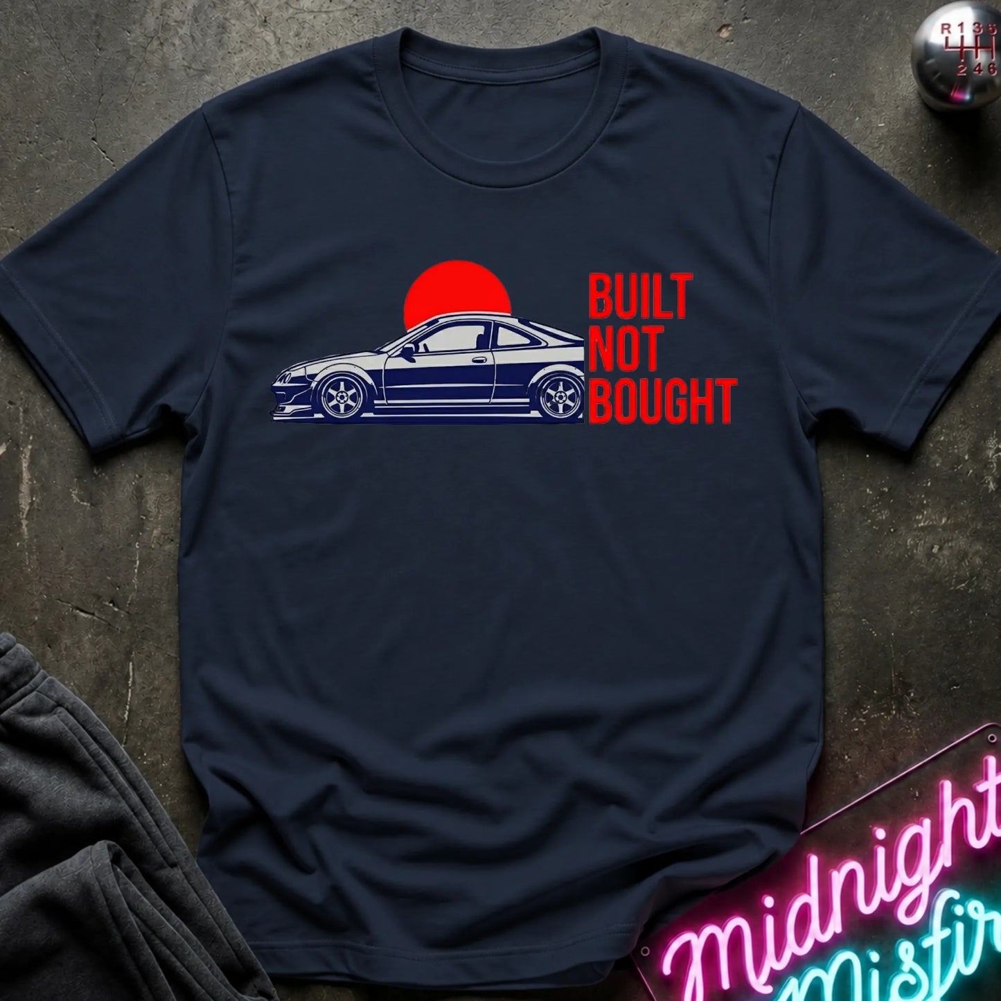 A navy blue graphic t-shirt laid flat on a dark textured surface. The shirt features a graphic of a blue sports coupe car in front of a red sun, with the text 'BUILT NOT BOUGHT' printed in red.