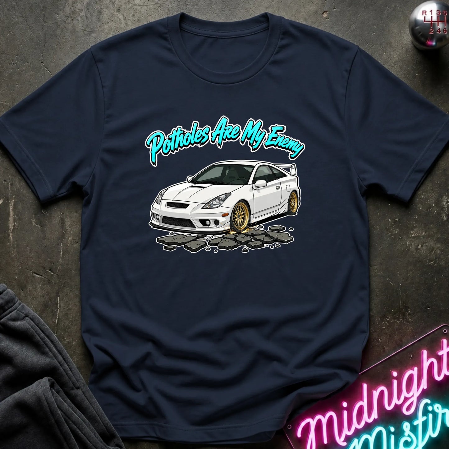 A navy blue graphic t-shirt laid flat on a dark textured surface. The shirt features a white sports car with gold wheels and the text 'Potholes Are My Enemy' in a teal cursive font.