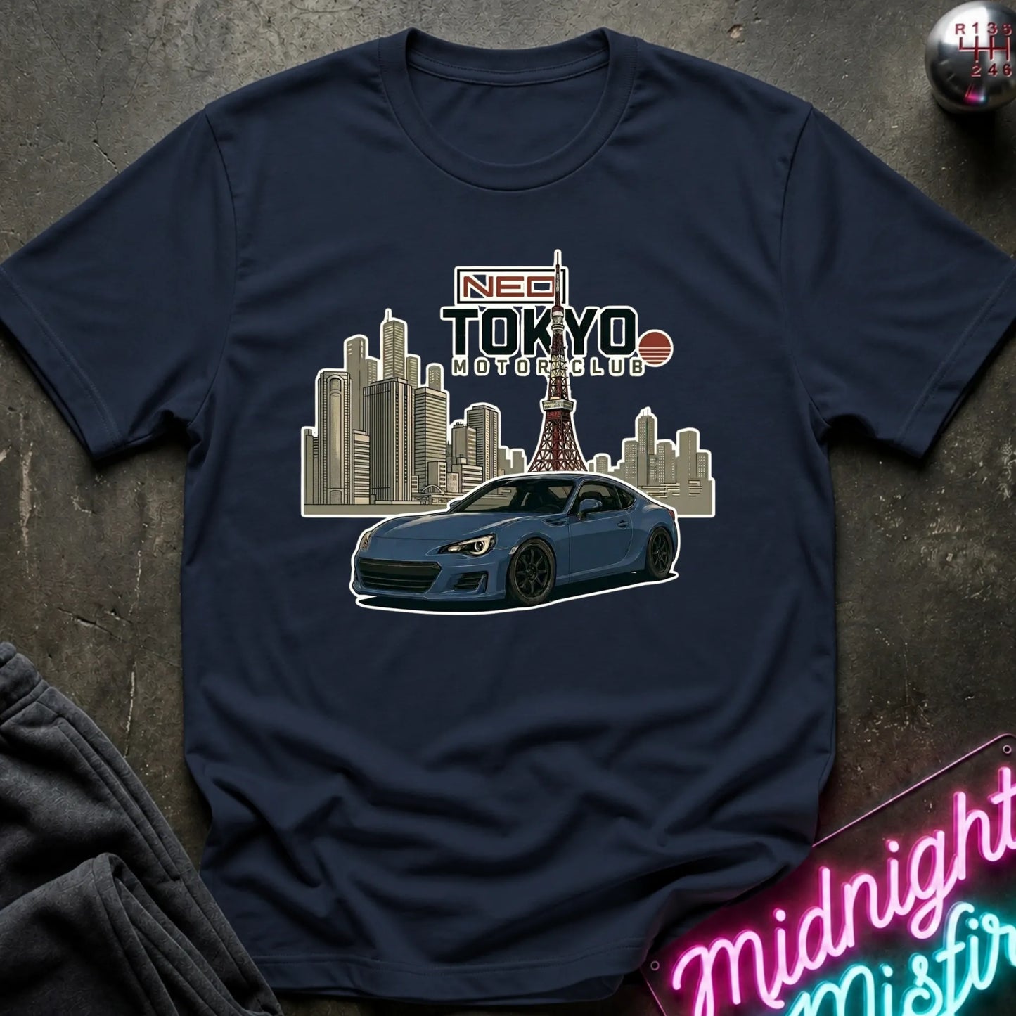 A navy short-sleeve graphic T-shirt laid flat on a dark textured surface. The shirt features a printed design of a blue sports car in front of a Tokyo city skyline with the Tokyo Tower, and text reading 'NEO TOKYO MOTORCLUB'.