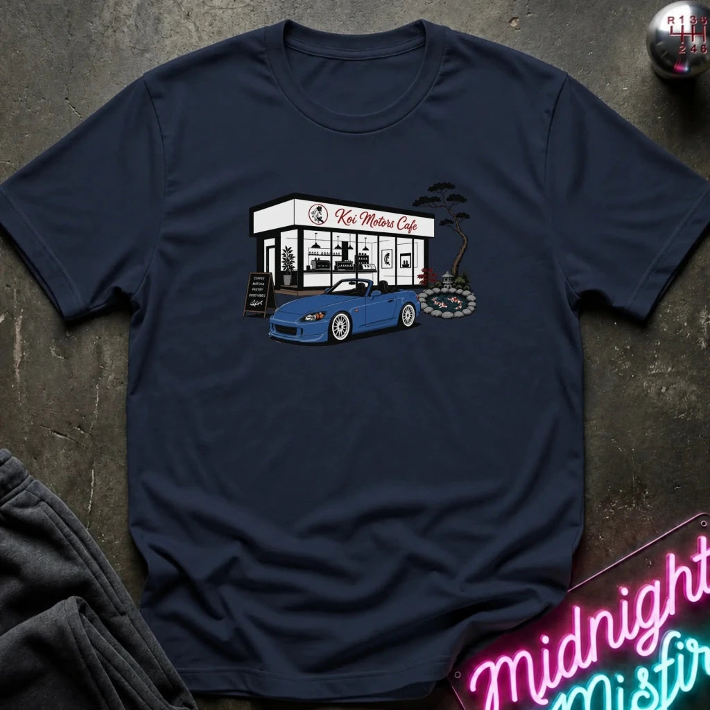 A navy blue short-sleeve t-shirt laid flat on a dark textured surface, featuring a graphic print of a blue sports car parked in front of a white cafe with the text 'Koi Motor Cafe'.