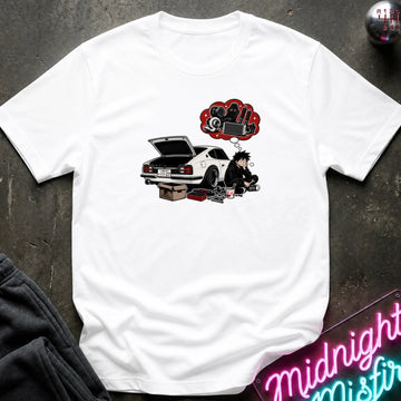 A white short-sleeve graphic t-shirt laid flat on a dark textured surface. The graphic features a white sports car with its trunk open, a cartoon character sitting beside it, and a red thought bubble filled with car parts.