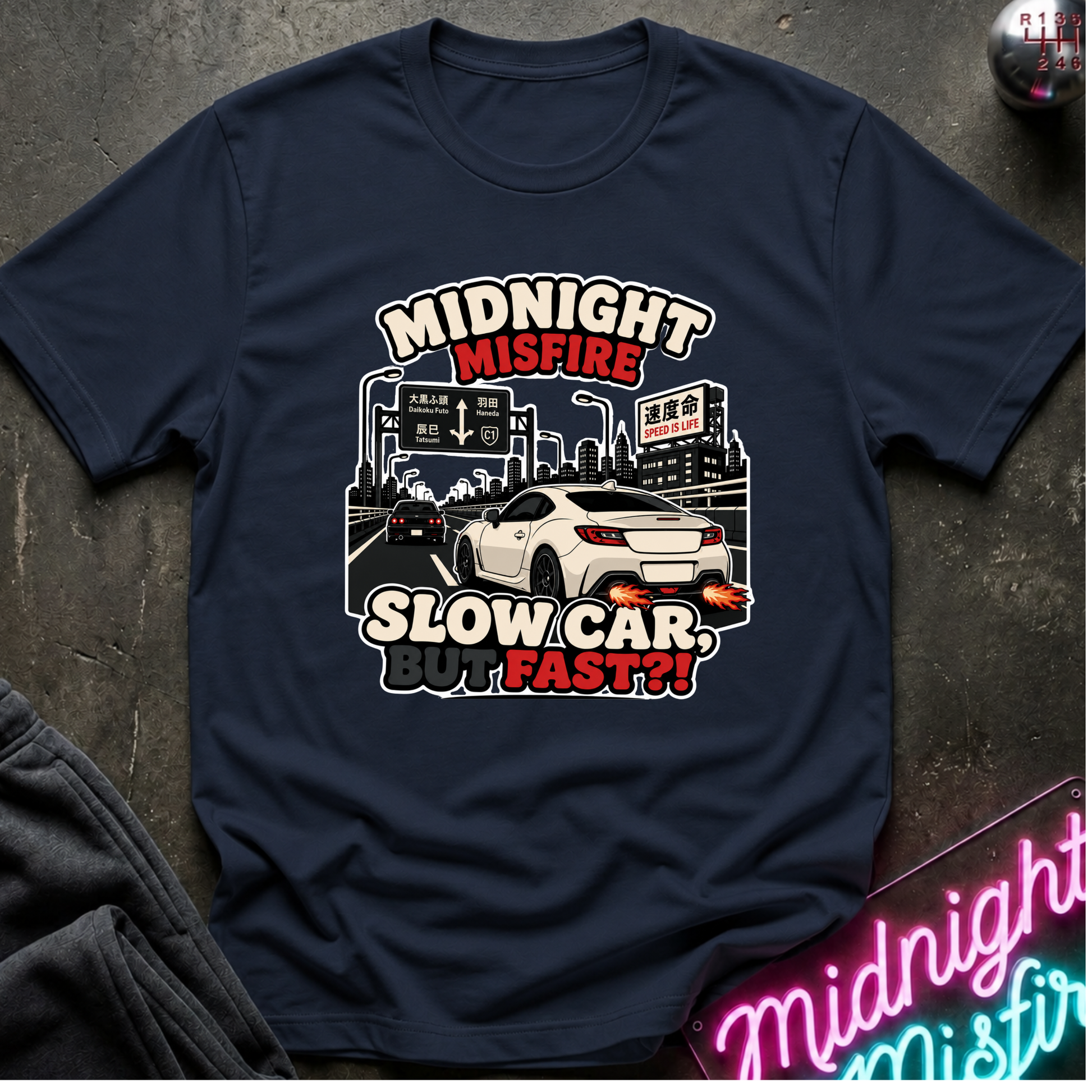 A navy blue graphic t-shirt laid flat on a dark textured surface. The shirt features a graphic of a white sports car driving towards city signs, with the text 'MIDNIGHT MISFIRE' and 'SLOW CAR.