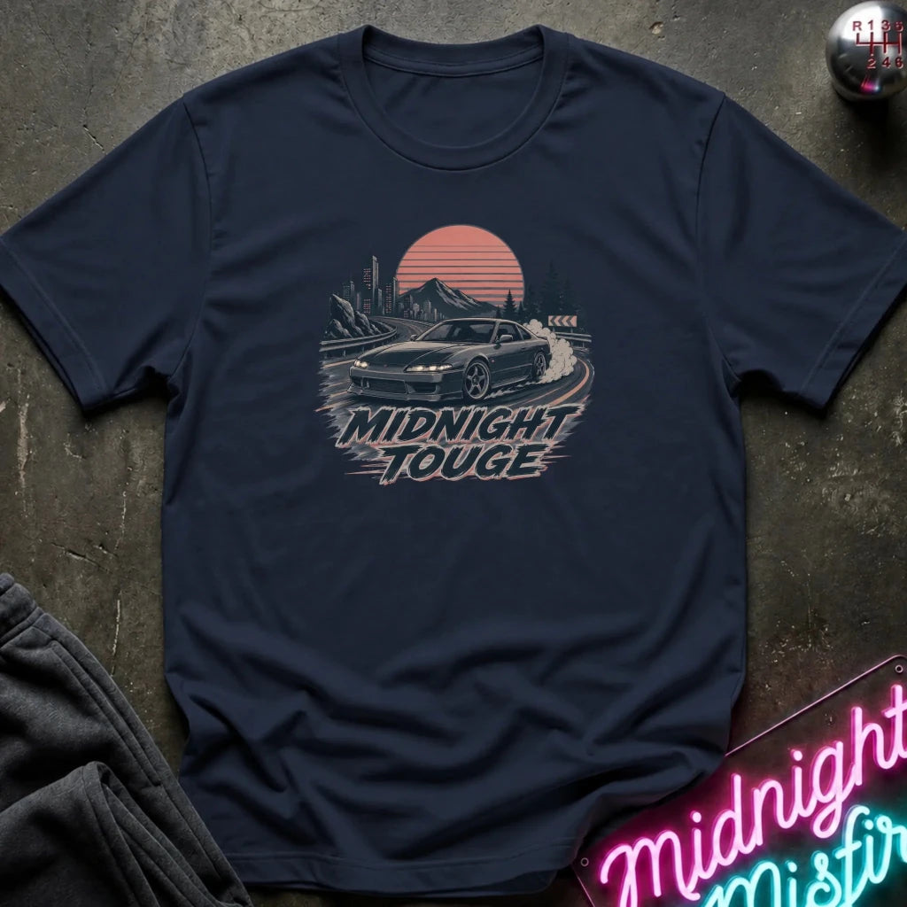 A navy blue graphic t-shirt laid flat on a dark textured surface. The shirt features a retro-style graphic of a sports car drifting in front of a sunset, with the text 'MIDNIGHT TOUGHE' printed below.
