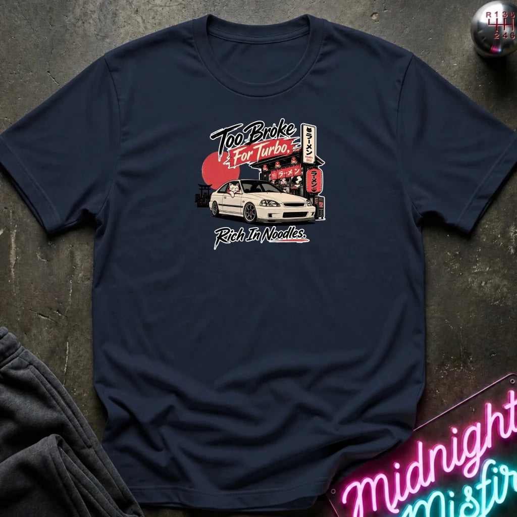 A navy blue graphic t-shirt laid flat on a dark textured surface. The shirt features a vintage-style chest graphic of a white sports car in front of a red sun and Japanese-style signs, with the text 'Too Broke For Turbo' and 'Rich In Noodles'.