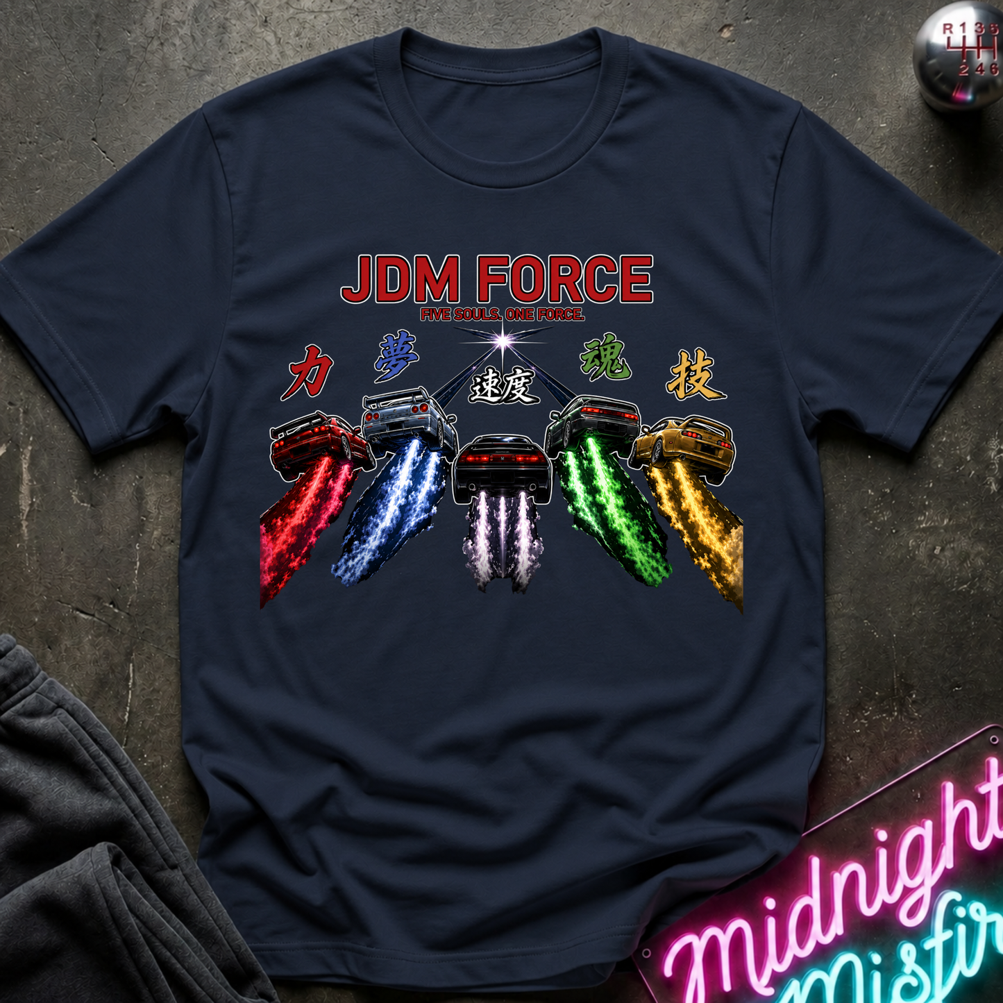 A navy blue graphic t-shirt laid flat on a dark textured surface. The shirt features a colorful graphic of cars with red, blue, white, green, and gold trails, with the text 'JDM FORCE'.