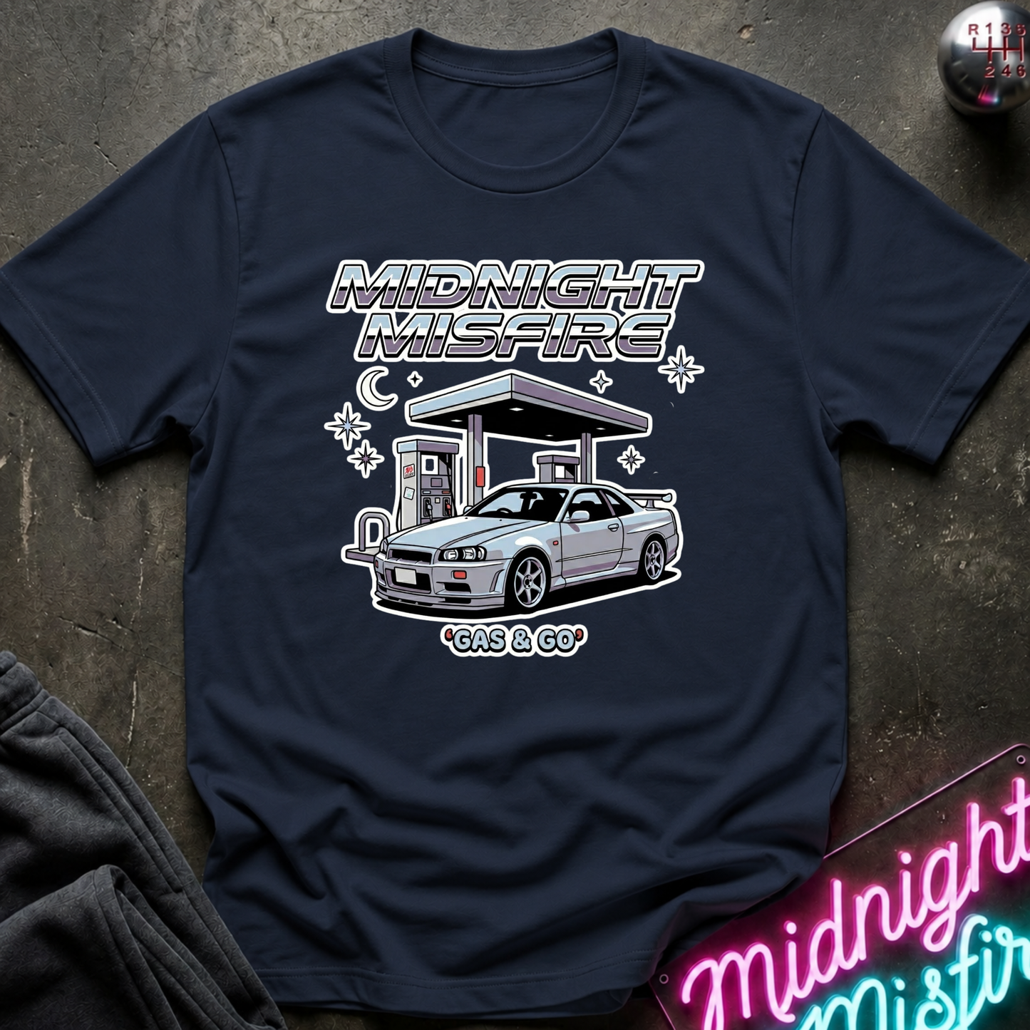 A navy blue graphic t-shirt laid flat on a dark textured surface. The shirt features a white and silver graphic of a Nissan Skyline R34 sports car at a gas station under the text 'MIDNIGHT MISFIRE' and 'GAS GO'.