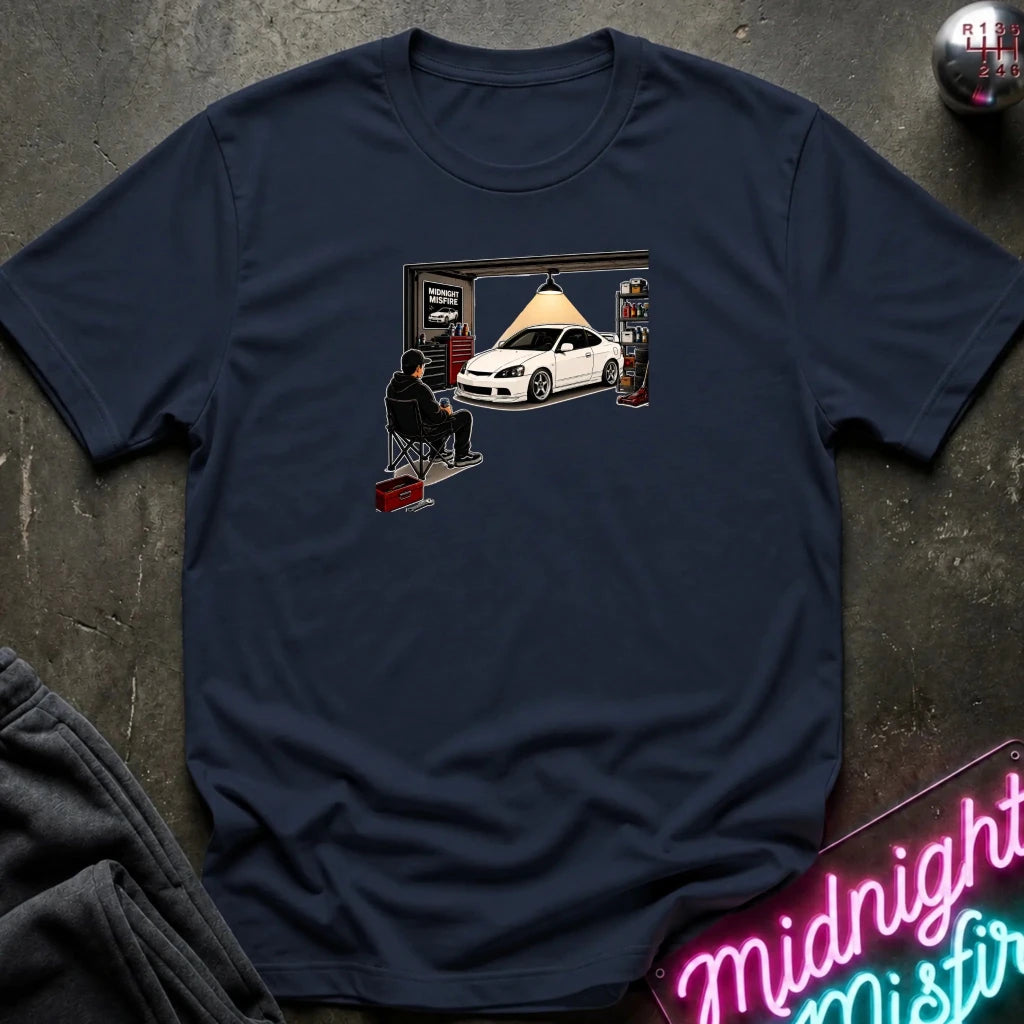 A navy blue short-sleeve t-shirt with a graphic print of a white car in a garage, laid flat on a dark textured surface with a neon sign and folded sweatpants nearby.