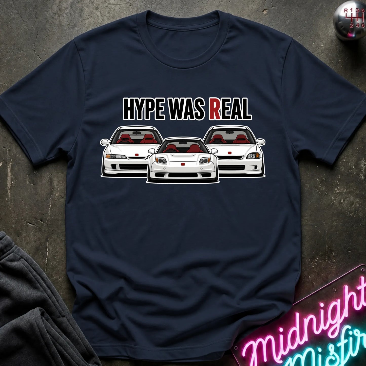 A navy blue short-sleeve graphic t-shirt laid flat on a dark textured surface. The shirt features a white and red graphic print of three sports cars with the text 'HYPE WAS REAL' above them, with 'R' highlighted in red.