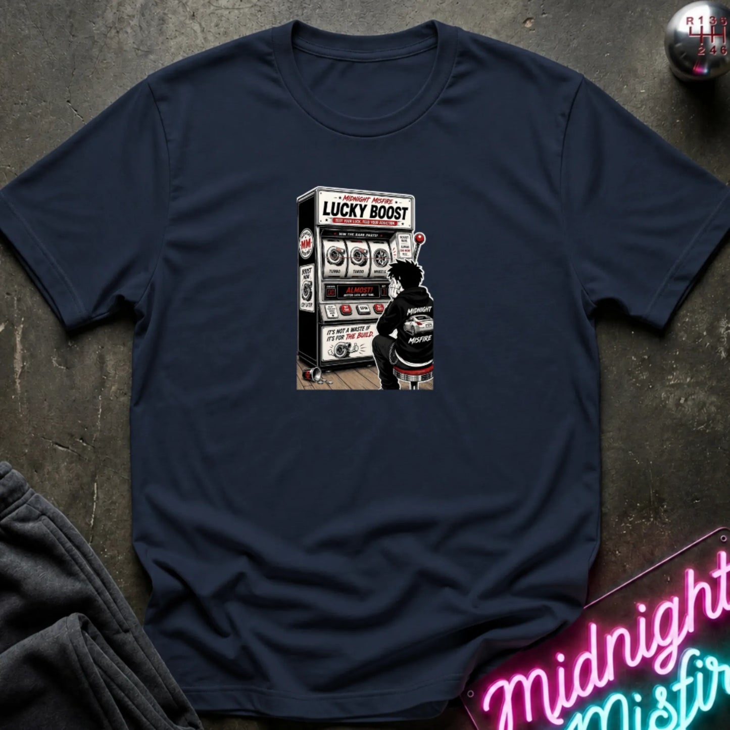 A navy  short-sleeve t-shirt with a retro-style graphic of a slot machine, displayed flat on a dark textured surface with a neon sign and sweatpants partially visible.