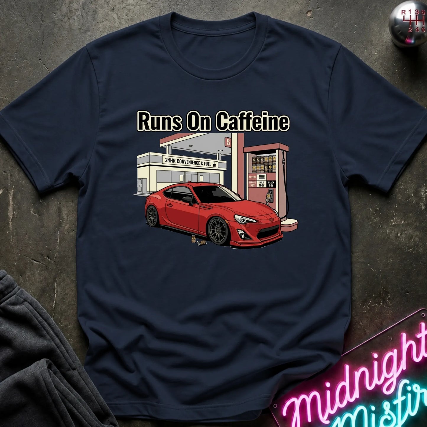 navy short-sleeve graphic T-shirt laid flat on a dark textured surface. The shirt features a printed illustration of a red sports car parked in front of a convenience store and gas station, with the text 'Runs On Caffeine' above the graphic.