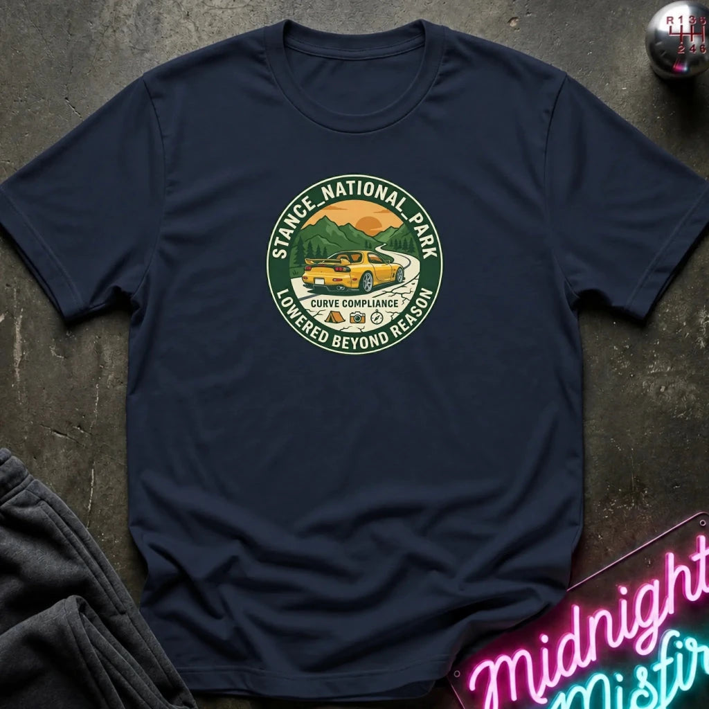 A navy blue crewneck t-shirt laid flat on a dark textured surface. The center of the shirt features a circular graphic print for 'Stance National Park' with a yellow car, mountains, and the text 'Lowered Beyond Reason'.
