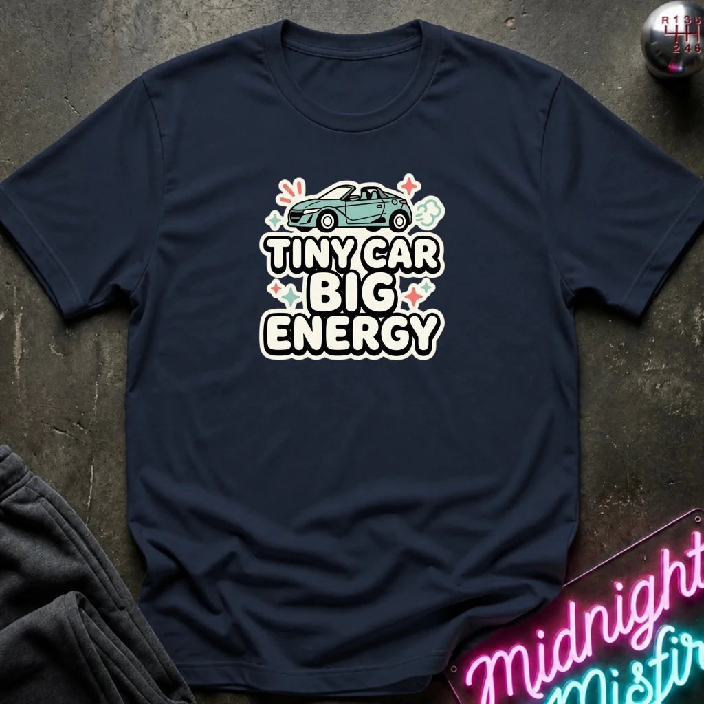 A navy blue graphic t-shirt laid flat on a dark textured surface. The shirt features a graphic of a small teal car above the text 'TINY CAR BIG ENERGY' in white and pink lettering.