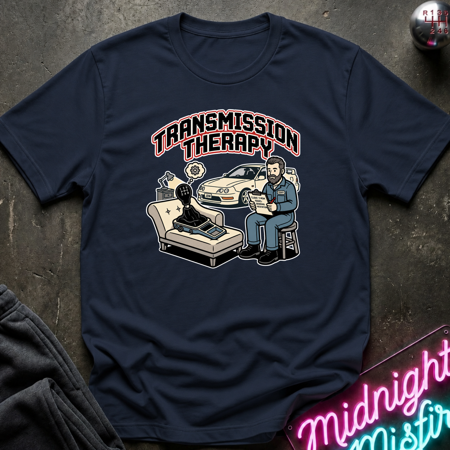 A navy blue graphic t-shirt laid flat on a dark textured surface. The shirt features a retro-style illustration of a therapist sitting next to a car with the text 'TRANSMISSION THERAPY' above it.