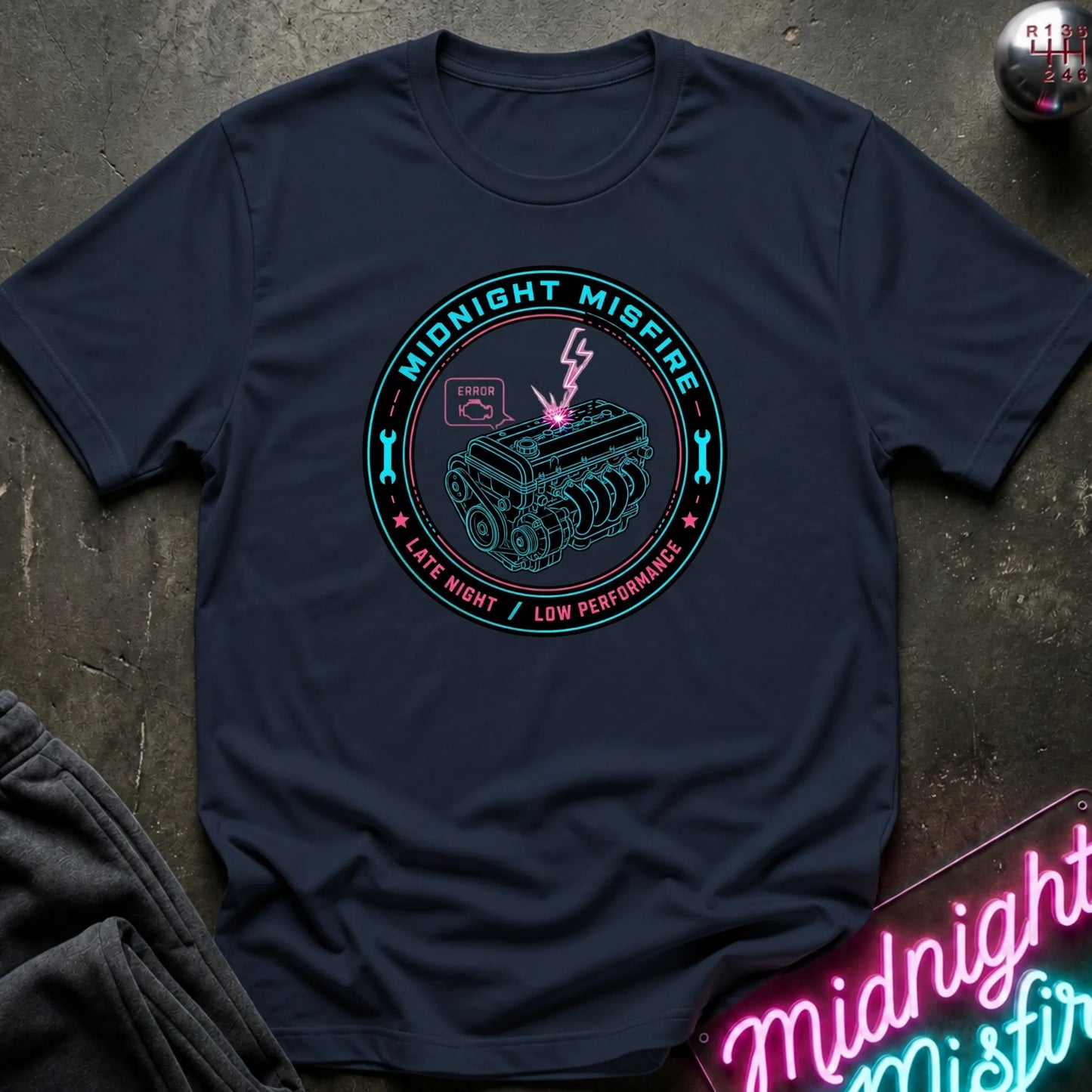 A navy blue crewneck t-shirt laid flat on a dark textured surface, featuring a circular graphic with a teal engine illustration, pink lightning bolt, and the words 'MIDNIGHT MISFIRE' and 'LATE NIGHT / LOW PERFORMANCE'. A matching neon-style sign is partially visible in the bottom right corner.