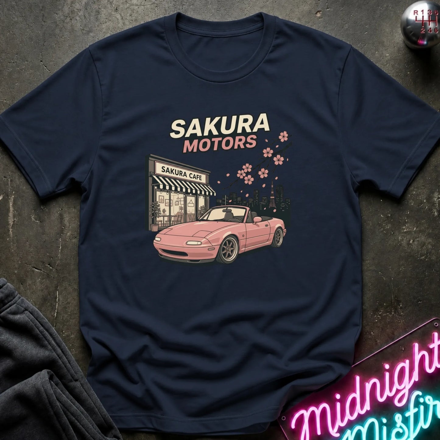 A navy blue graphic t-shirt laid flat on a dark textured surface. The shirt features a retro-style print of a pink convertible sports car in front of a 'Sakura Motors' cafe, with cream and pink text and cherry blossoms.