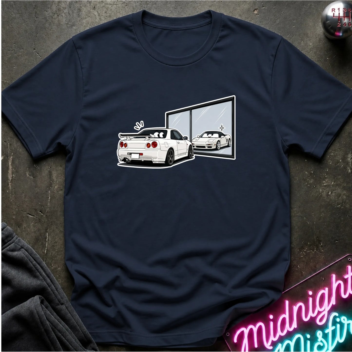 A navy blue short-sleeve t-shirt laid flat on a dark textured surface, featuring a graphic print of a white sports car and its reflection in a window. A neon sign  is visible in the bottom right corner.