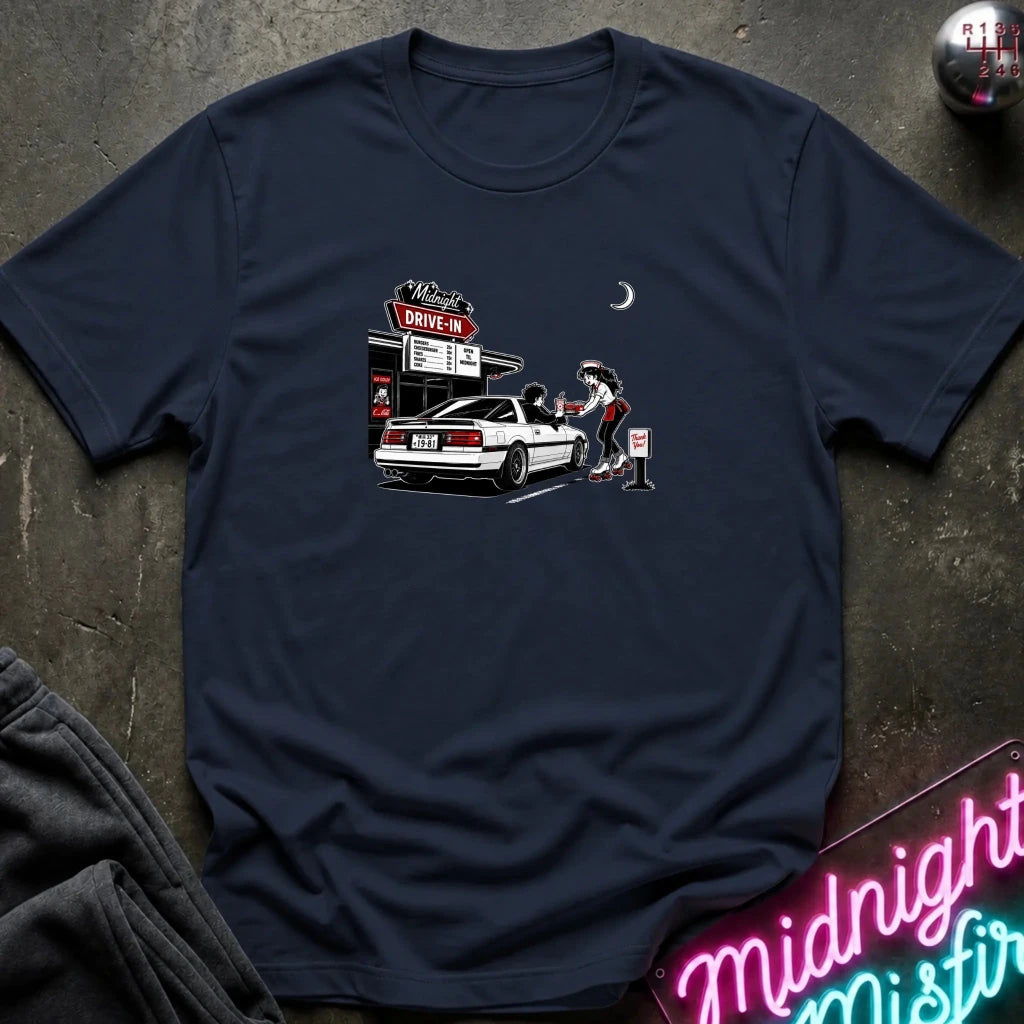 A navy short-sleeve t-shirt laid flat on a dark textured surface, featuring a graphic print of a woman pumping gas at a drive-in station next to a white car. 