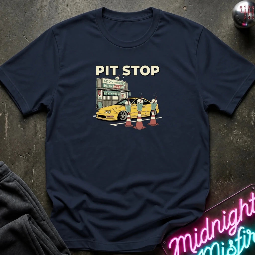A navy blue graphic t-shirt laid flat on a dark textured surface. The shirt features a retro-style illustration of a yellow sports car at a pit stop with the text 'PIT STOP' above it.