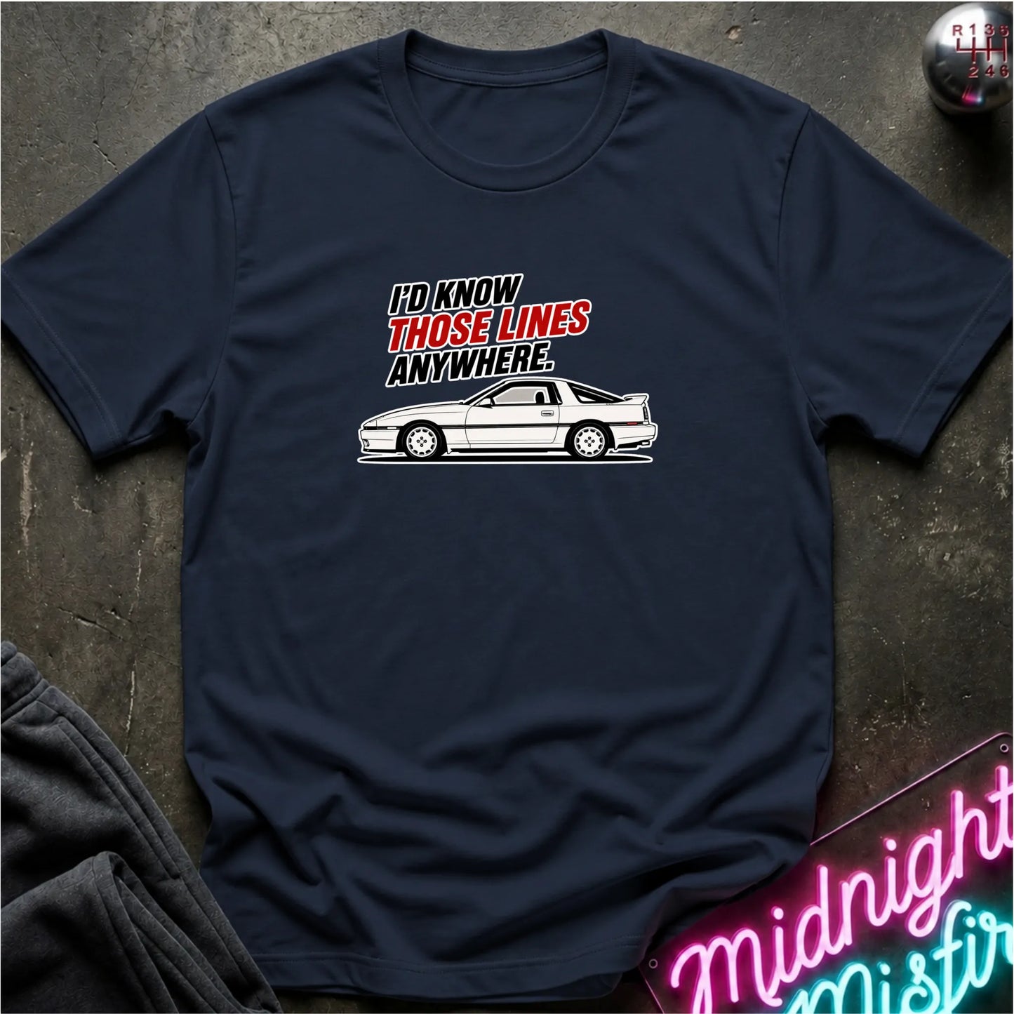 A navy blue graphic t-shirt laid flat on a dark textured surface. The shirt features a white line art graphic of a sports car and the text 'I'D KNOW THOSE LINES ANYWHERE.