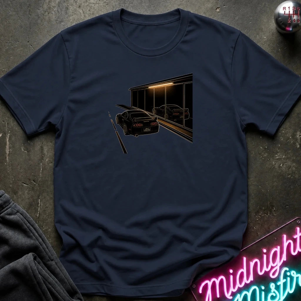 A navy blue short-sleeve t-shirt laid flat on a dark textured surface, featuring a graphic print of a black sports car in front of a dark building with warm light. 