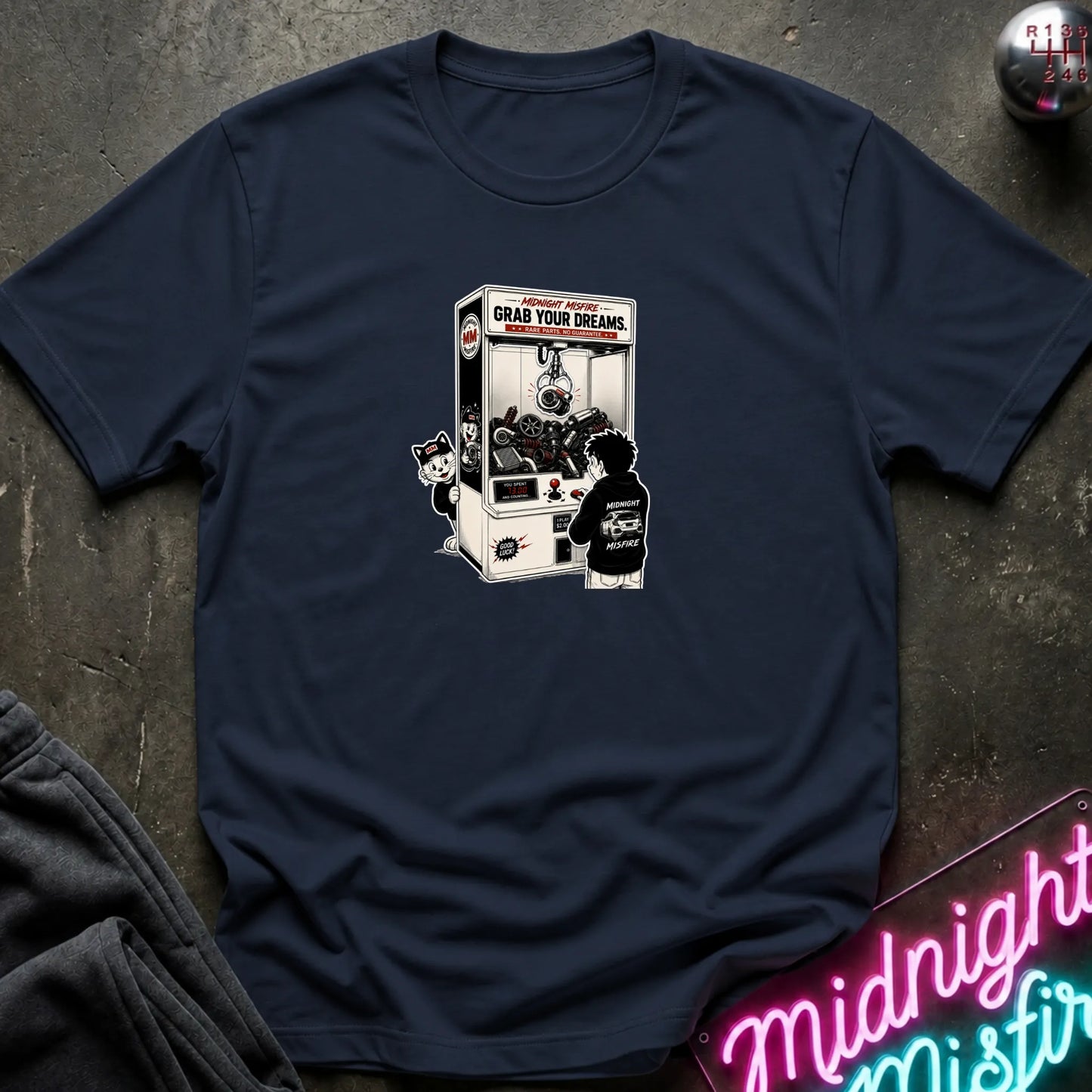 A navy blue short-sleeve crewneck t-shirt laid flat on a dark textured surface, featuring a central graphic of a person in a black hoodie playing a claw machine. 