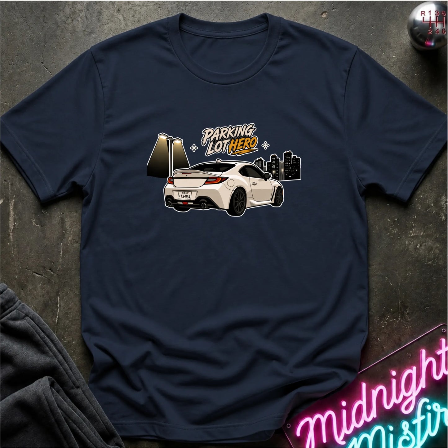 A navy blue crewneck t-shirt laid flat on a dark textured surface. The shirt features a graphic print of a white sports car with the text 'PARKING LOT HERO' in stylized lettering, alongside a gold-toned gate and a black city skyline.