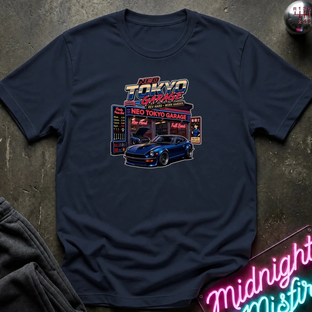 A navy blue graphic t-shirt laid flat on a dark textured surface. The shirt features a colorful retro-style 'Neo Tokyo Garage' print with a blue sports car and neon sign details.