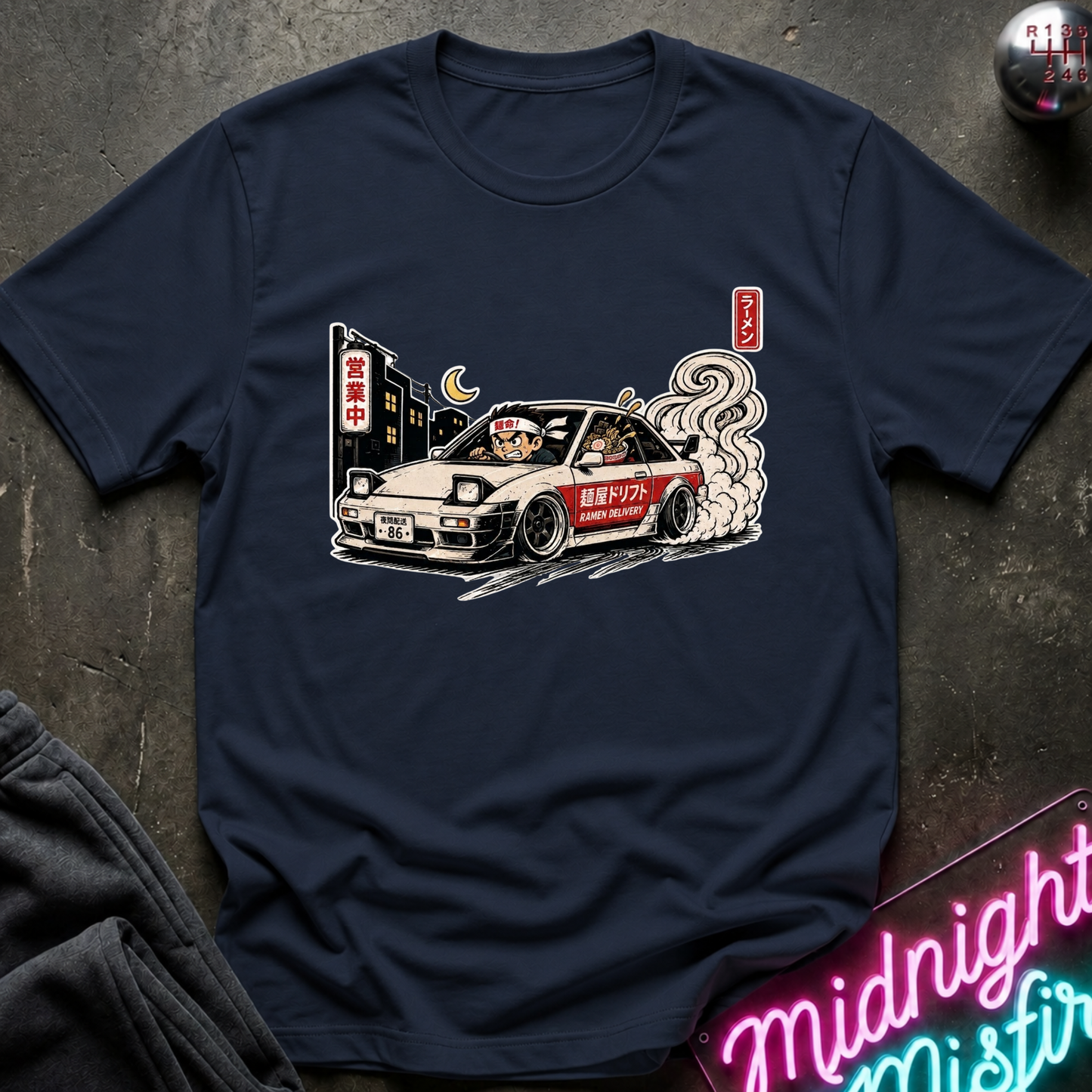 A navy blue short-sleeve t-shirt with a graphic print of a white and red sports car driving at night, styled flat on a dark textured surface with a neon sign and dark sweatpants partially visible.