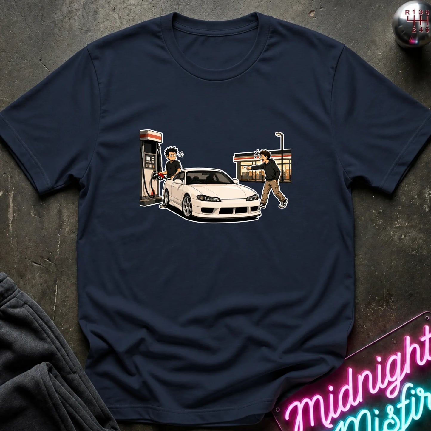 A navy blue short-sleeve t-shirt with a graphic print of a white sports car at a gas station, laid flat on a dark textured surface with a neon sign and dark fabric partially visible.