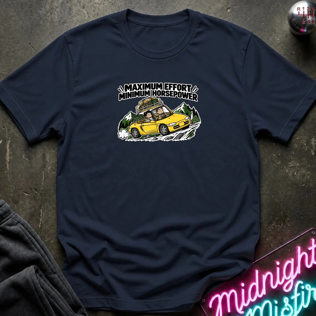 A navy blue graphic t-shirt laid flat on a dark textured surface. The graphic features a yellow sports car with a driver and passengers, with the text 'MAXIMUM EFFORT MINIMUM HORSEPOWER' above it.