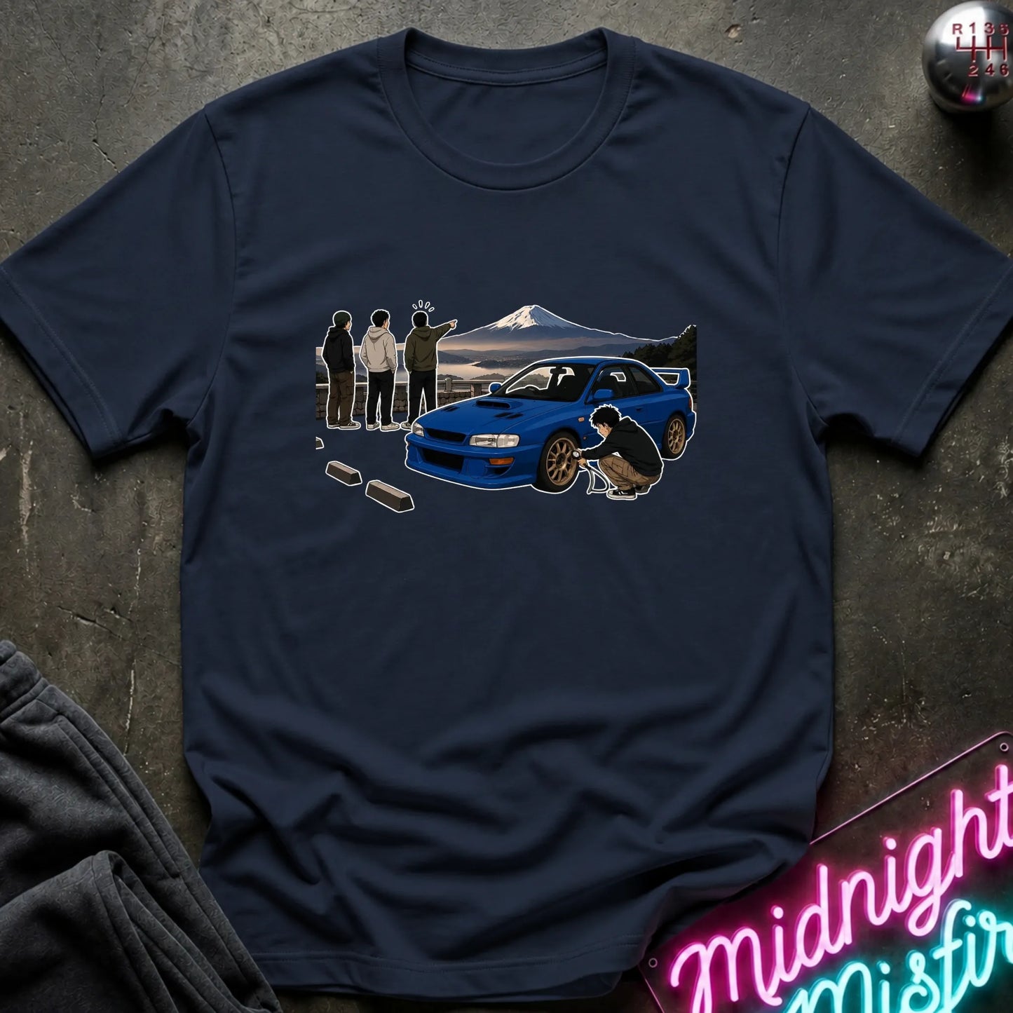 A navy blue short-sleeve t-shirt laid flat on a dark textured surface, featuring a graphic print of a blue sports car in front of a mountain with silhouettes of people and a driver crouching beside it. A neon sign and folded dark fabric are partially visible around the shirt.