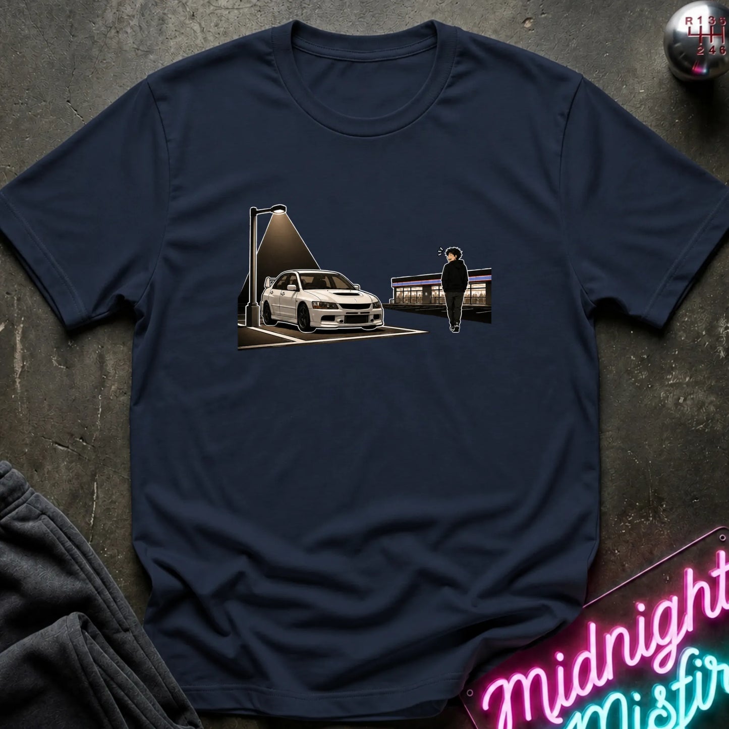 A navy blue short-sleeve t-shirt laid flat on a dark textured surface, featuring a central graphic print of a white sports car under a streetlamp with a silhouette figure standing nearby. A neon sign is partially visible in the bottom right corner.
