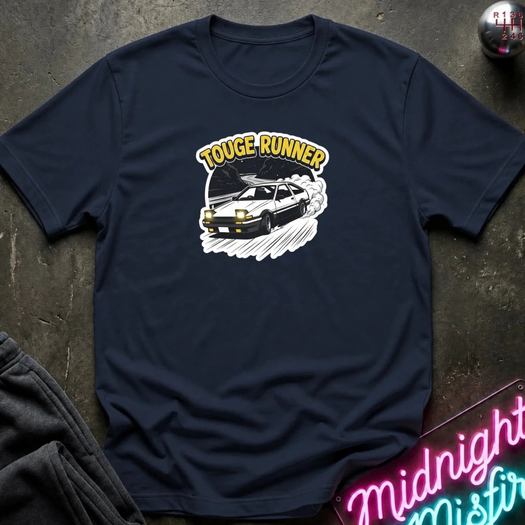 navy short-sleeve t-shirt laid flat on a dark textured surface, featuring a graphic of a black sports car with the words 'TOUGE RUNNER' in yellow above it. A neon sign and folded dark fabric are partially visible around the edges.