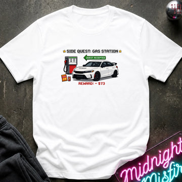 A white short-sleeve graphic t-shirt laid flat on a dark textured surface. The graphic features a white sports car, a red gas pump, and text that reads 'SIDE QUEST: GAS STATION' and 'REWARD: $73'.