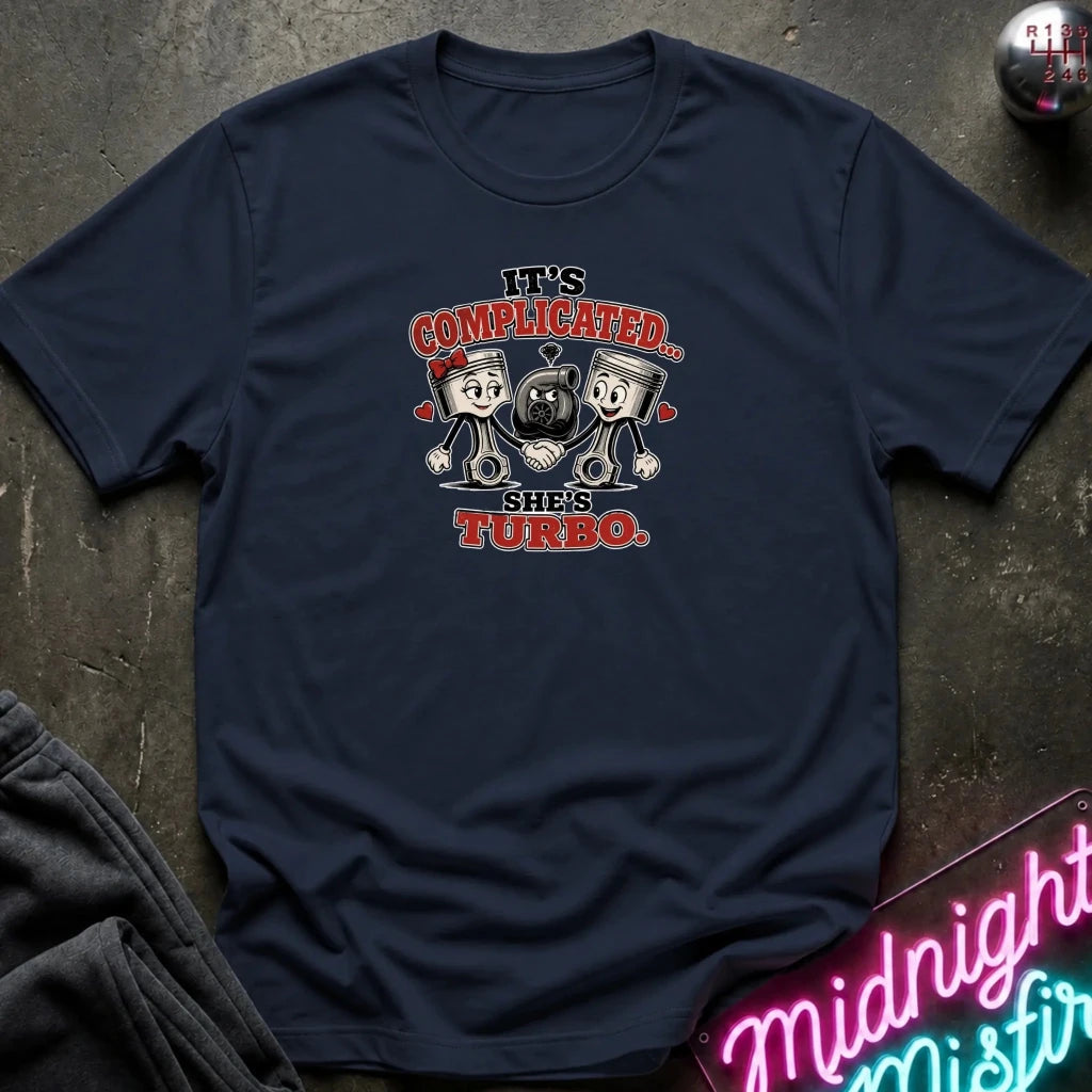 navy short-sleeve graphic T-shirt laid flat on a dark textured surface. The shirt features a cartoon graphic of two pistons holding a heart-shaped turbocharger, with the text 'IT'S COMPLICATED.